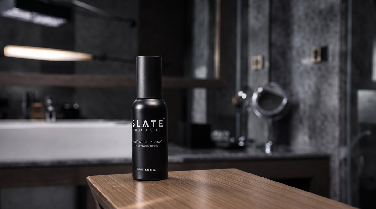 A matte black bottle of SLATE PROJECT Hair Reset Spray standing on a wooden table in a modern, dark-toned bathroom setting.