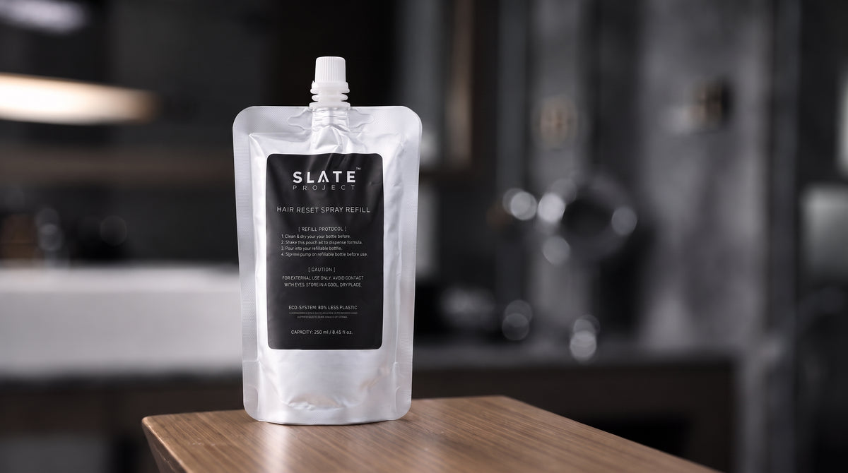 A silver 250 ml SLATE Hair Reset Spray Refill pouch with a black label, standing on a wooden surface in a dark interior setting.