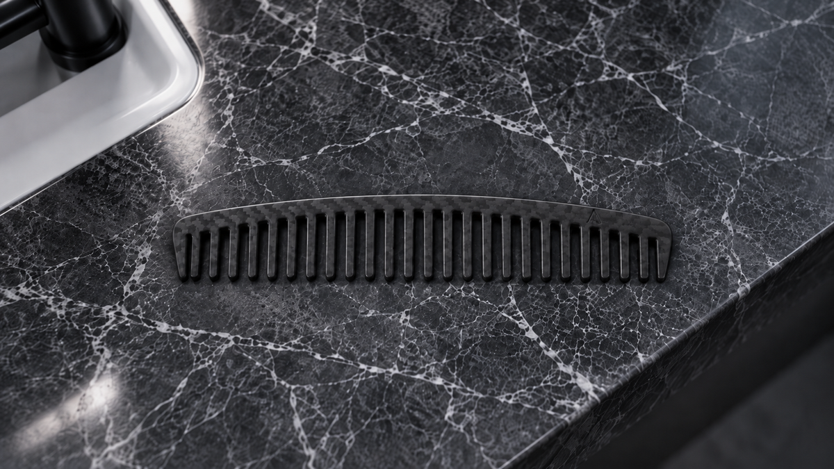 A black curved carbon fiber SLATE PROJECT hair comb with wide teeth, resting on a dark gray and white marble countertop.